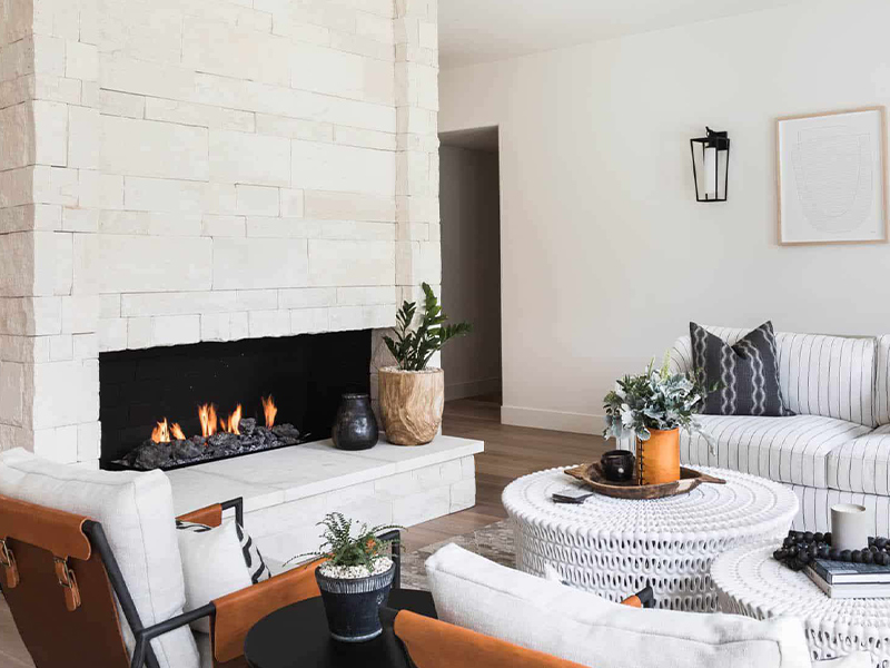 Cozy living room with a white stone fireplace featuring a lit fire and a black interior. The room includes a striped sofa with patterned cushions, a pair of white woven coffee tables, and chairs with leather straps. Decorative plants and vases add a touch of greenery, and a wall-mounted lantern light fixture enhances the space's warm and inviting atmosphere.