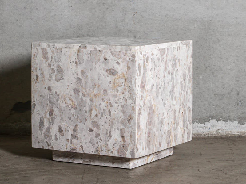 Square marble side table with a textured, light-colored surface, set against a plain grey background. The table has a sturdy, minimalist design, emphasizing the natural patterns and variations in the stone.