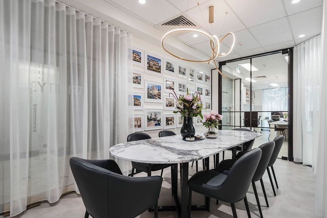 Elegant conference room with a long marble table surrounded by black chairs. The room features modern lighting fixtures, large windows with sheer curtains, and walls adorned with framed photographs. The setting is sophisticated and designed for professional meetings.