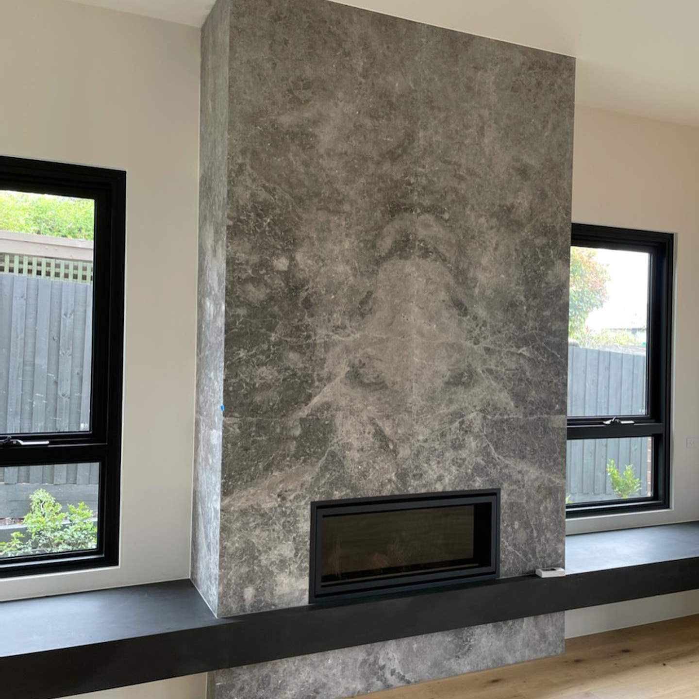 Modern fireplace with a tall, grey marble surround, flanked by two windows with black frames. The fireplace is set above a sleek black bench that runs along the length of the wall. The room has light wood flooring and a minimalist design, emphasizing clean lines and natural materials.